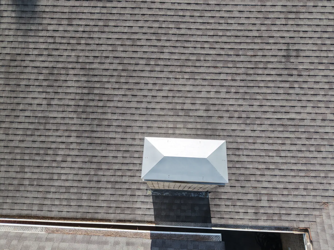 Overhead view of a chimney chase on an asphalt shingle roof fitted with an intact sloped metal chase cover