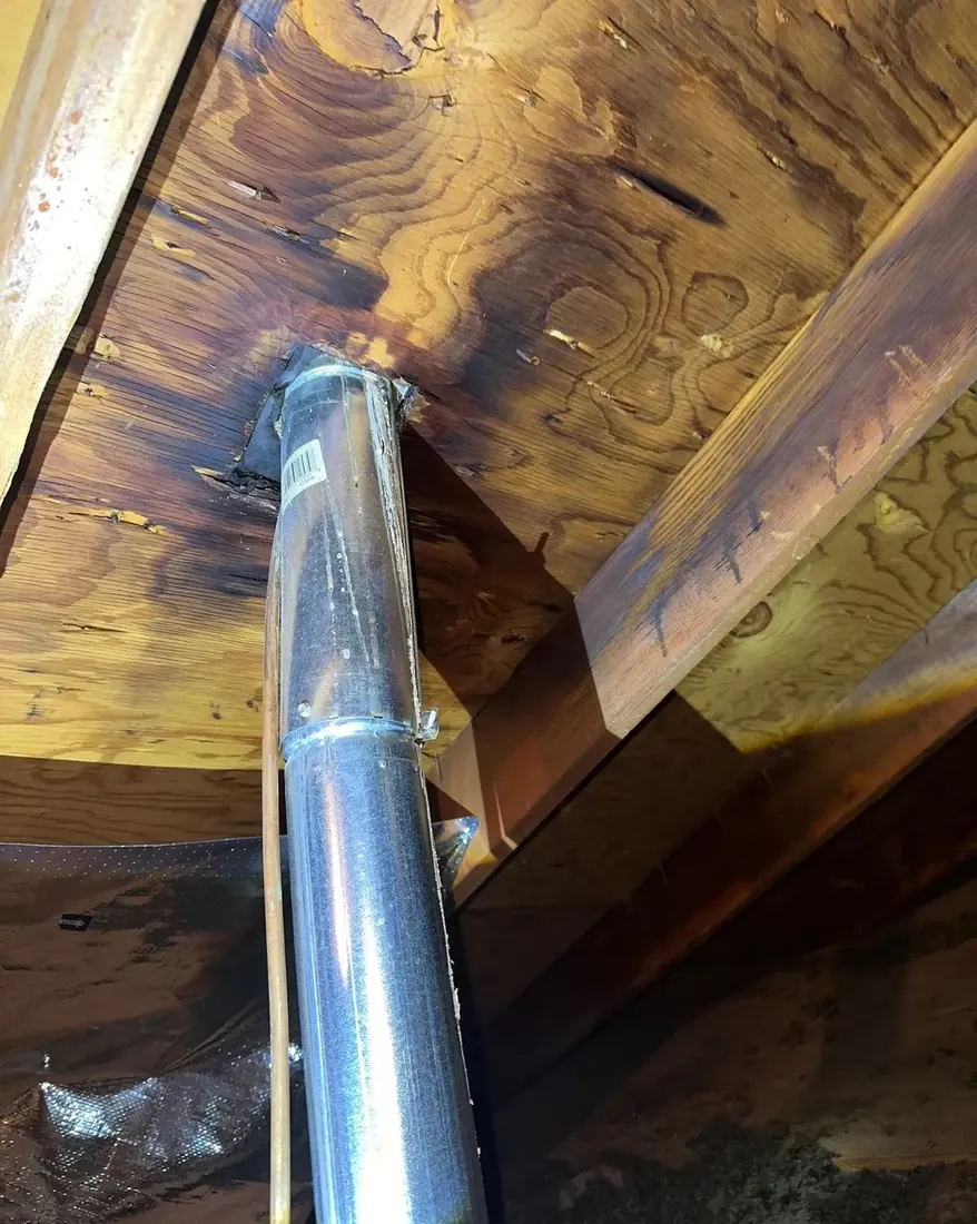 A metal flue pipe passing through roof decking in an attic with dark water staining on the wood around the penetration