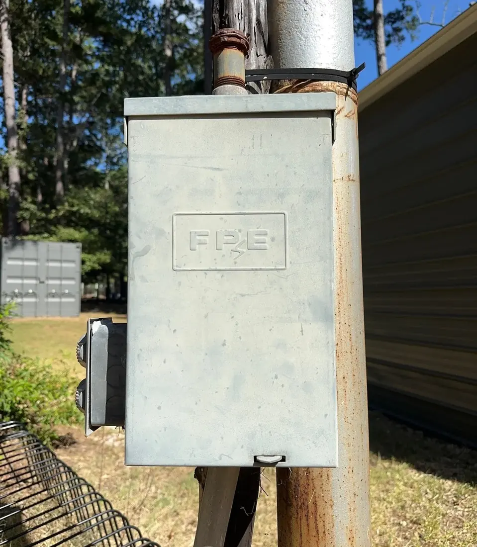 An exterior electrical disconnect enclosure on a pole with FPE embossed on the cover, weathered and rusting at the mounting pipe