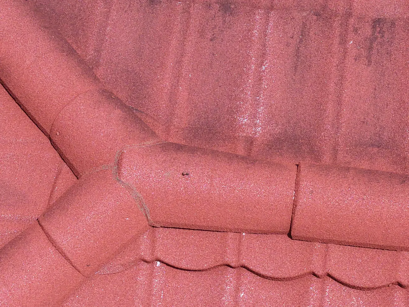 Close drone view of a red metal tile-profile roof showing a welded joint running along a panel seam below a hip