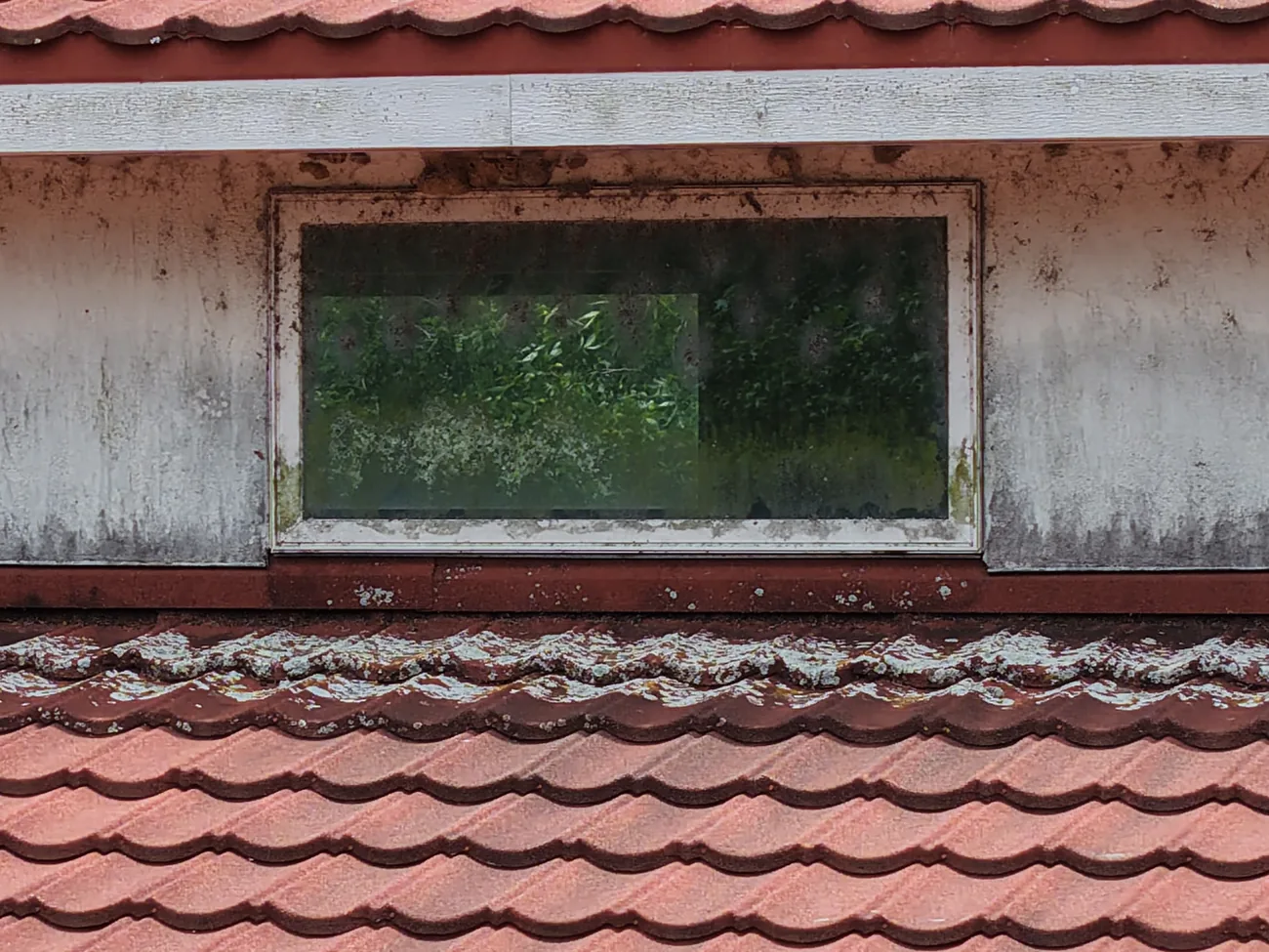 Drone view of a dormer window above a red metal roof with dark mildew and algae growth across the wall surface