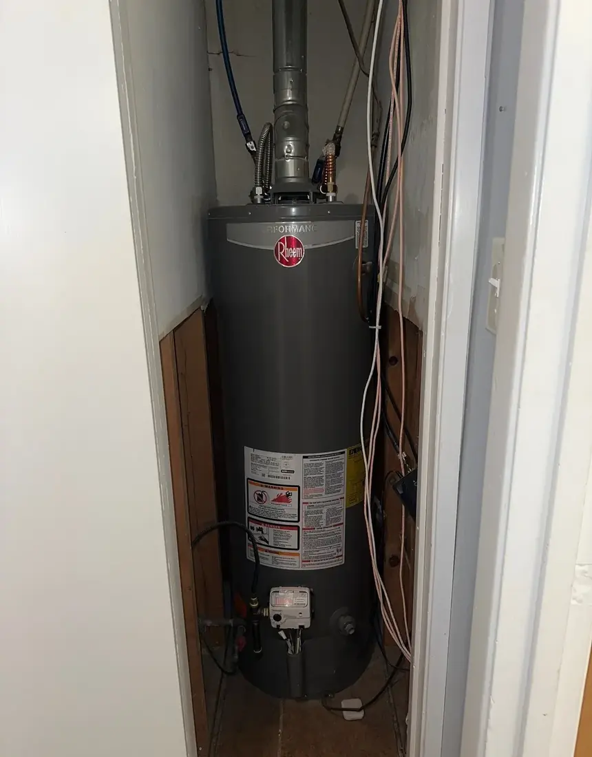 A Rheem gas water heater installed in a narrow interior closet with flue, gas line and water connections above