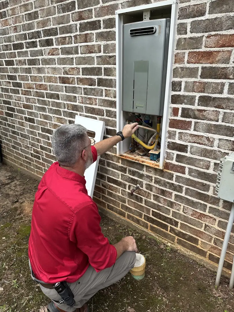 Jeff Stalnaker kneeling at an exterior brick wall with the cover removed from a tankless water heater, examining the gas and water connections inside