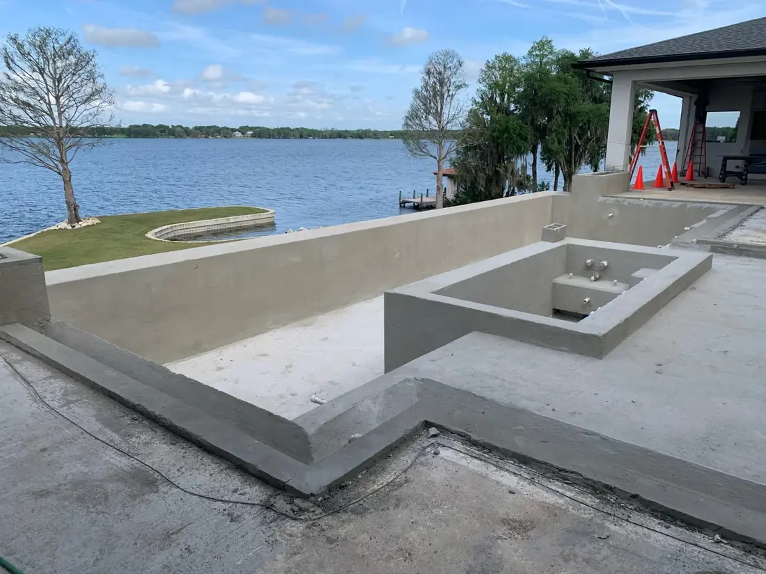 A residential pool shell under construction beside a lake, with the spa formed and plumbing stubbed